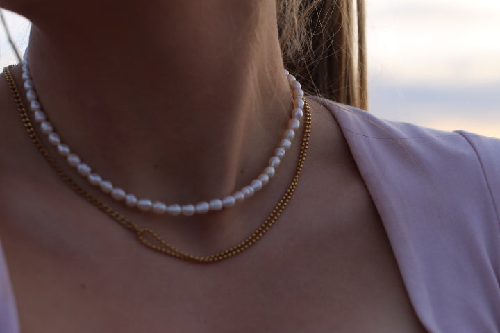 A close-up of a person's neck and upper chest wearing two layered necklaces: a white pearl necklace and a gold chain necklace, against a softly blurred outdoor background.